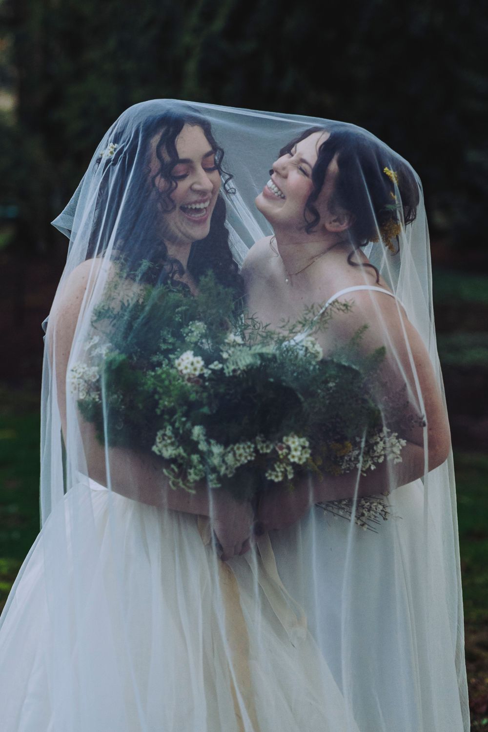 Two brides share a joyful moment together under a wedding veil while holding green and white floral bouquets.