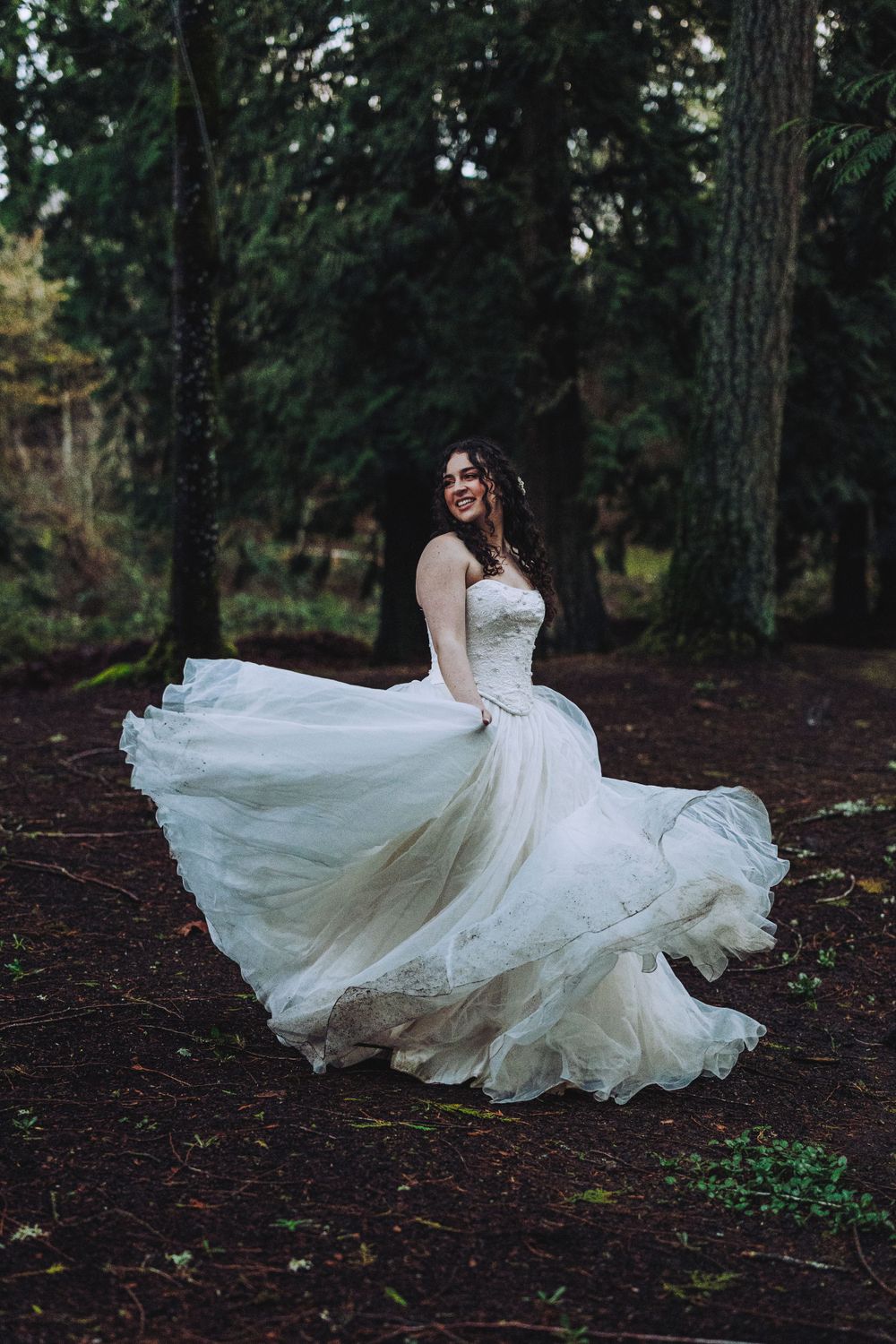 A bride in a flowing white dress twirls gracefully among tall pine trees in a moody forest setting.