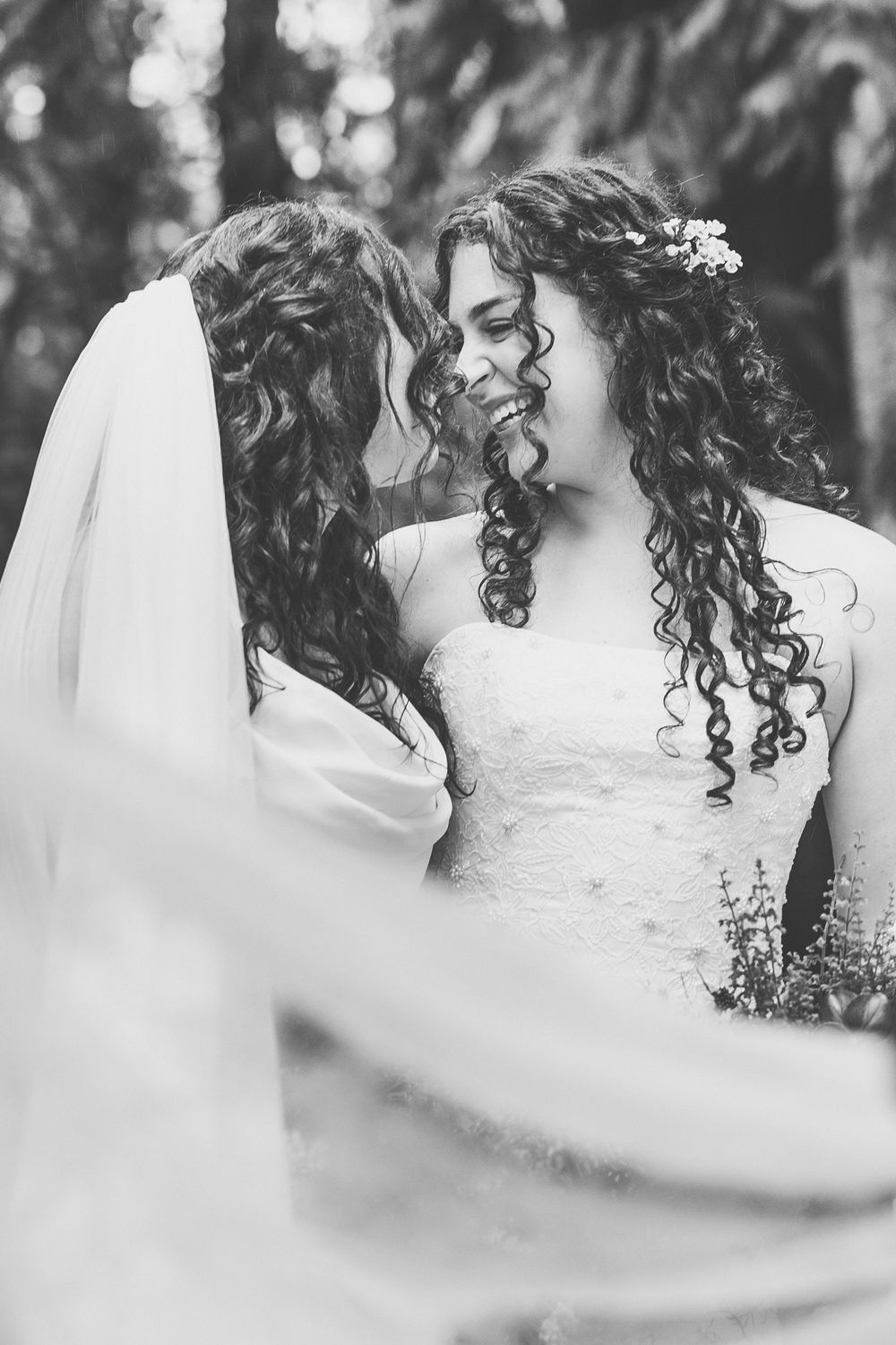 A romantic black and white wedding moment showing two brides with curly hair and floral accessories embracing.