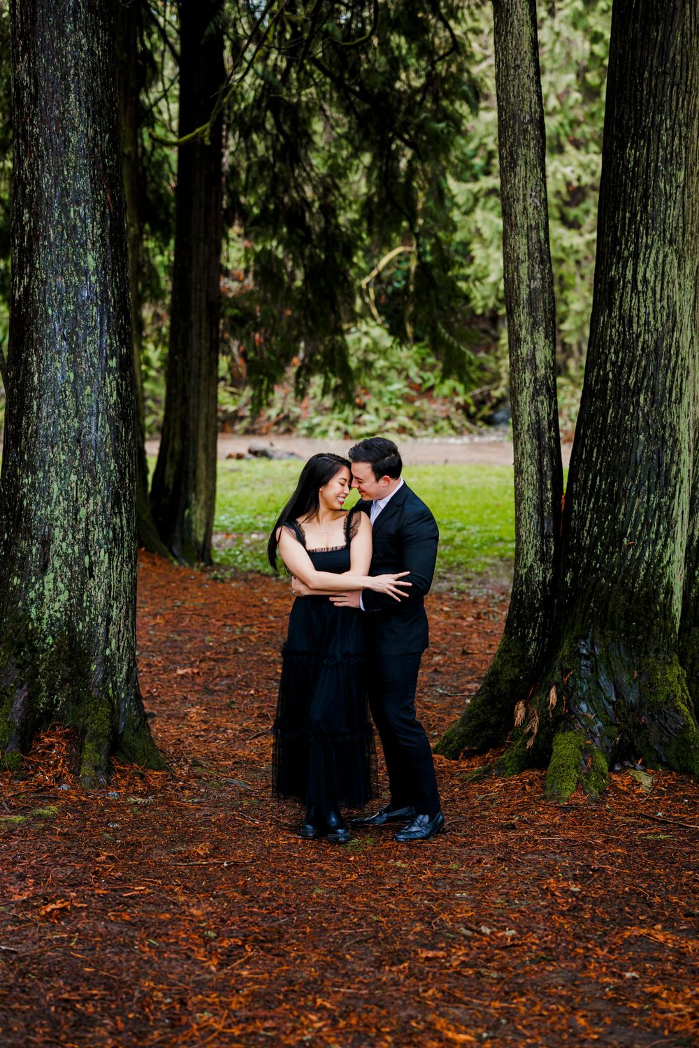A romantic couple sharing an intimate moment between tall evergreen trees on a forest path covered in fallen needles.