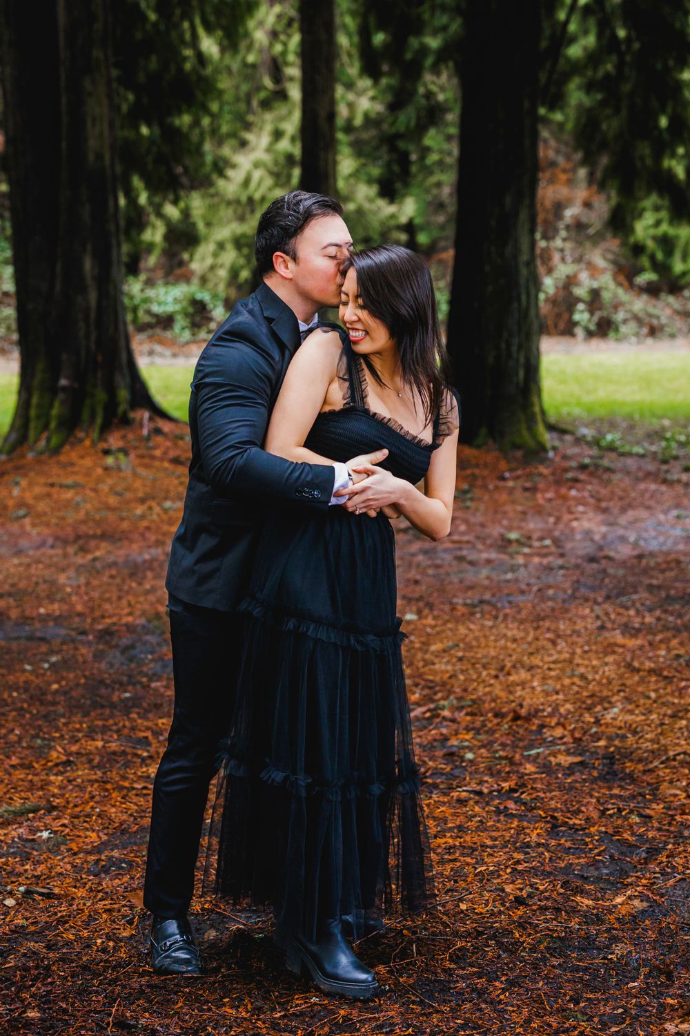 A couple in dark formal attire share an intimate moment while embracing on fallen autumn leaves in a forest setting.