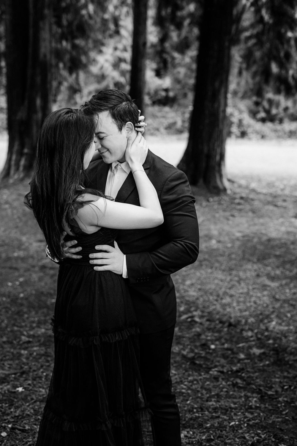 A romantic black and white photo of a couple embracing intimately among tall trees in a forest setting.