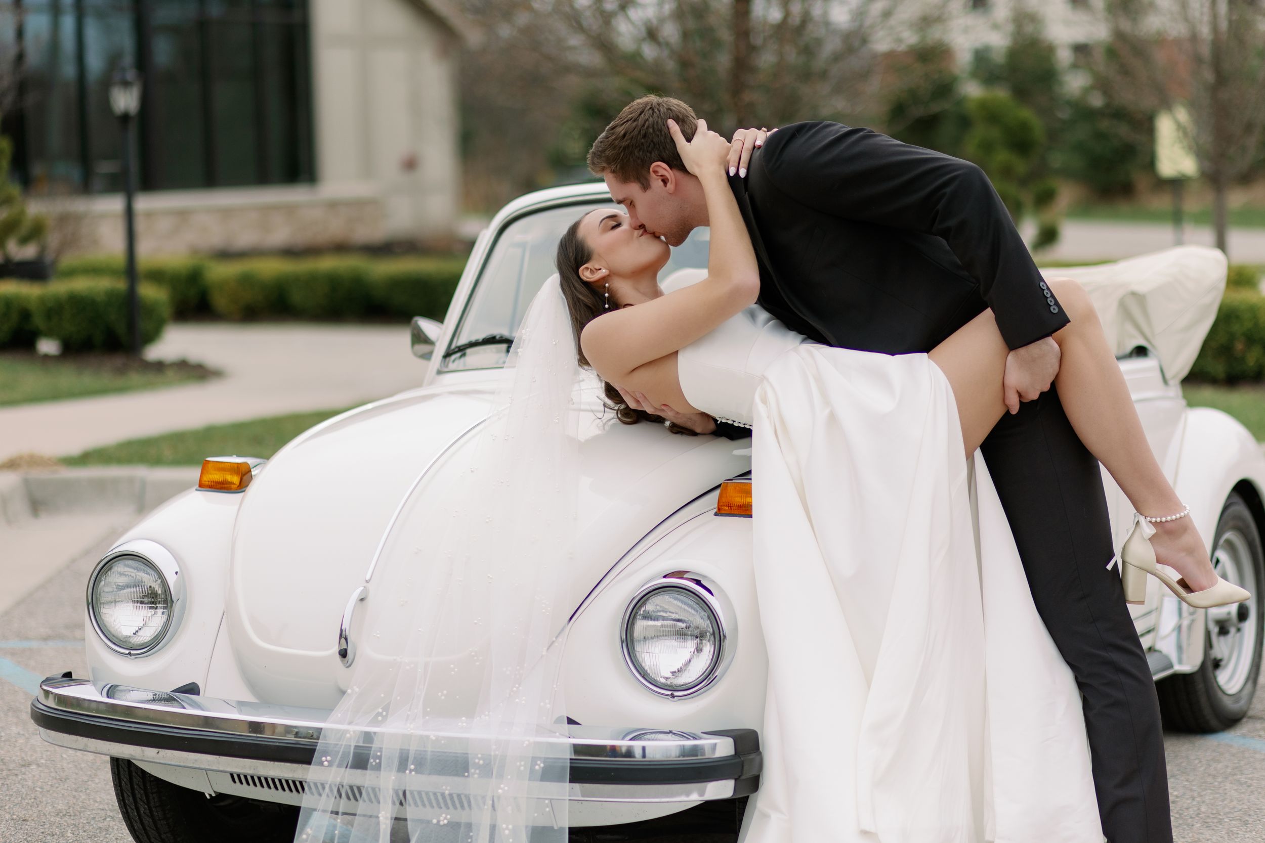 A couple shares a romantic moment while leaning against a classic white Volkswagen Beetle decorated for a wedding at Venue 3Two in GR.