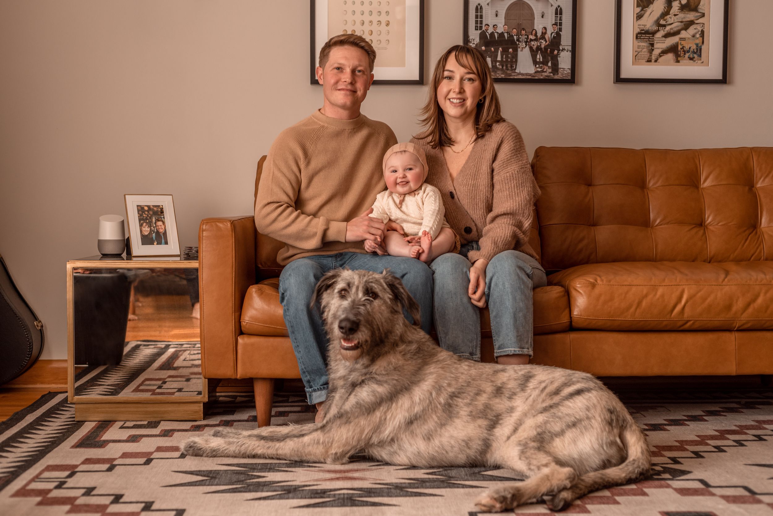 Family relaxes on brown leather couch with large gray dog on patterned rug.