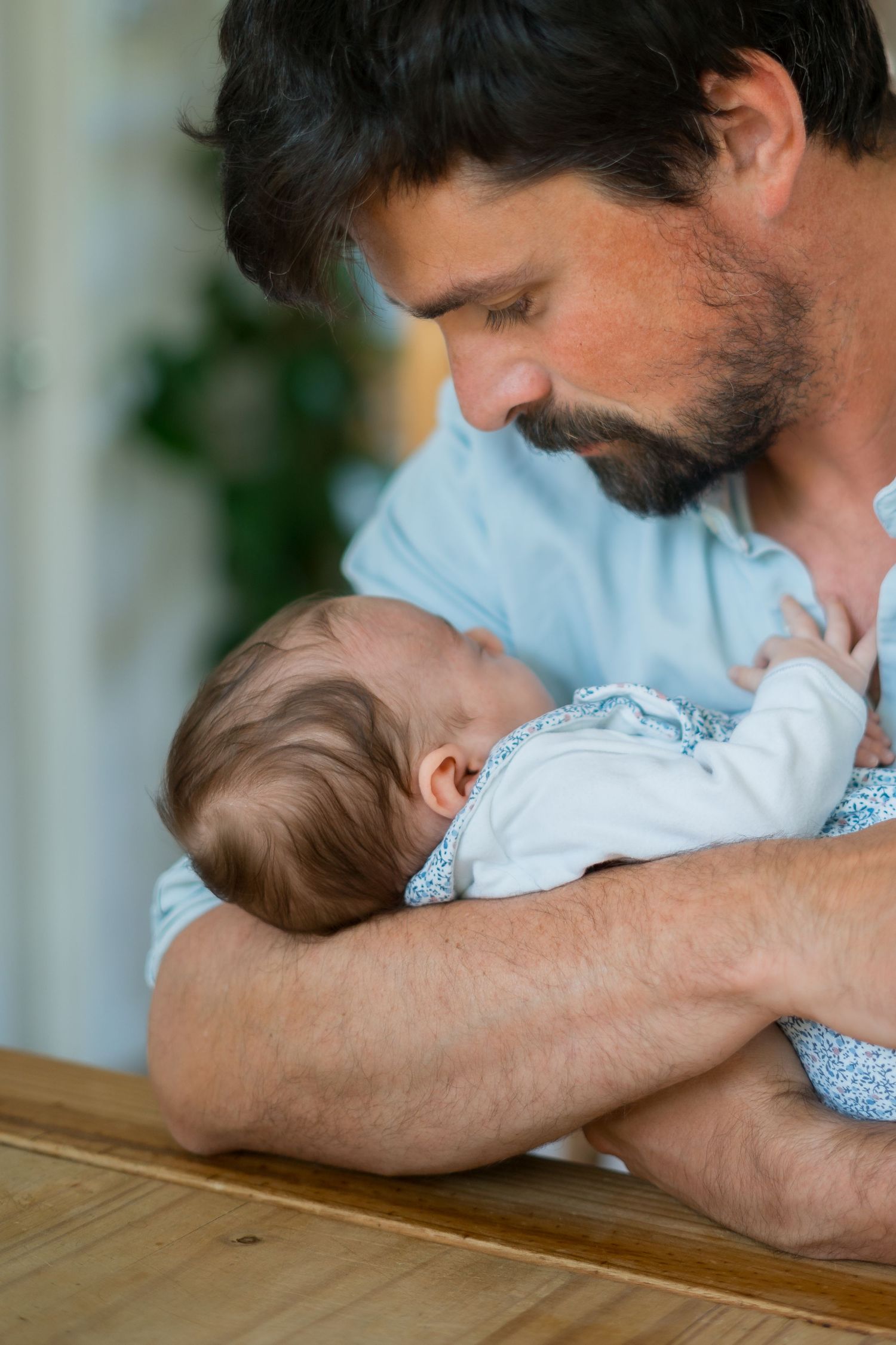 Bearded man in a light blue shirt tenderly holds sleeping baby against wooden table surface.