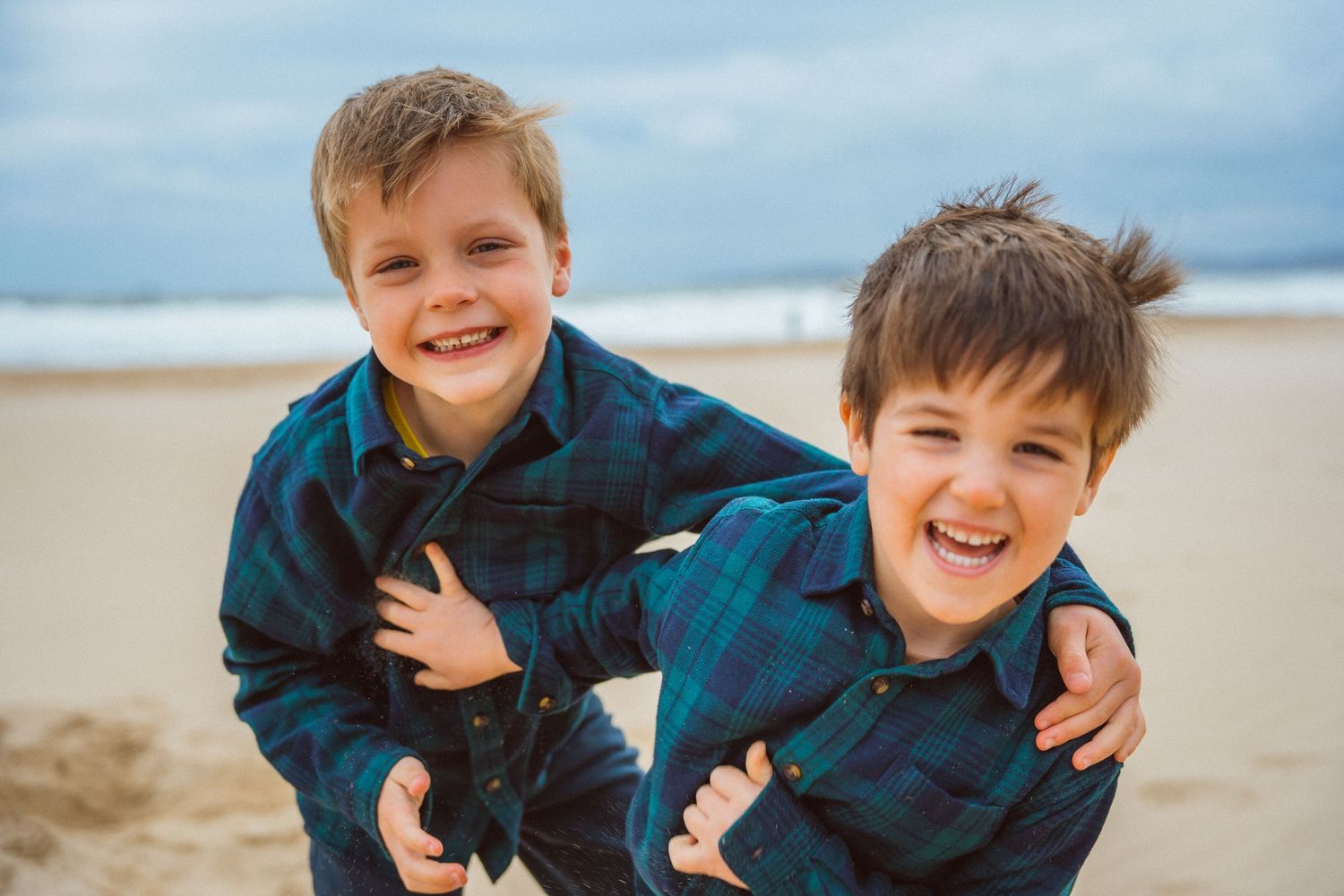 Two children in matching plaid shirts laugh together while playing on a sandy beach.