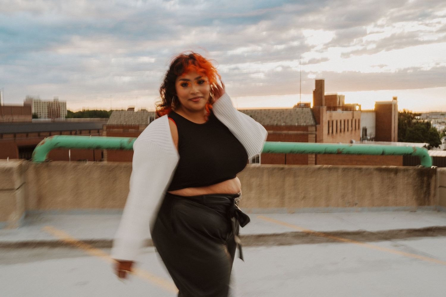 A person in a black crop top and white cardigan poses on an urban rooftop at sunset with city buildings in the background.