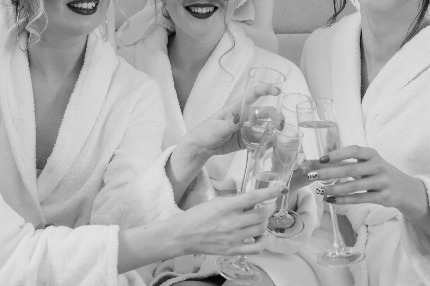 Group in white bathrobes smiling and toasting with champagne glasses in black and white photography.