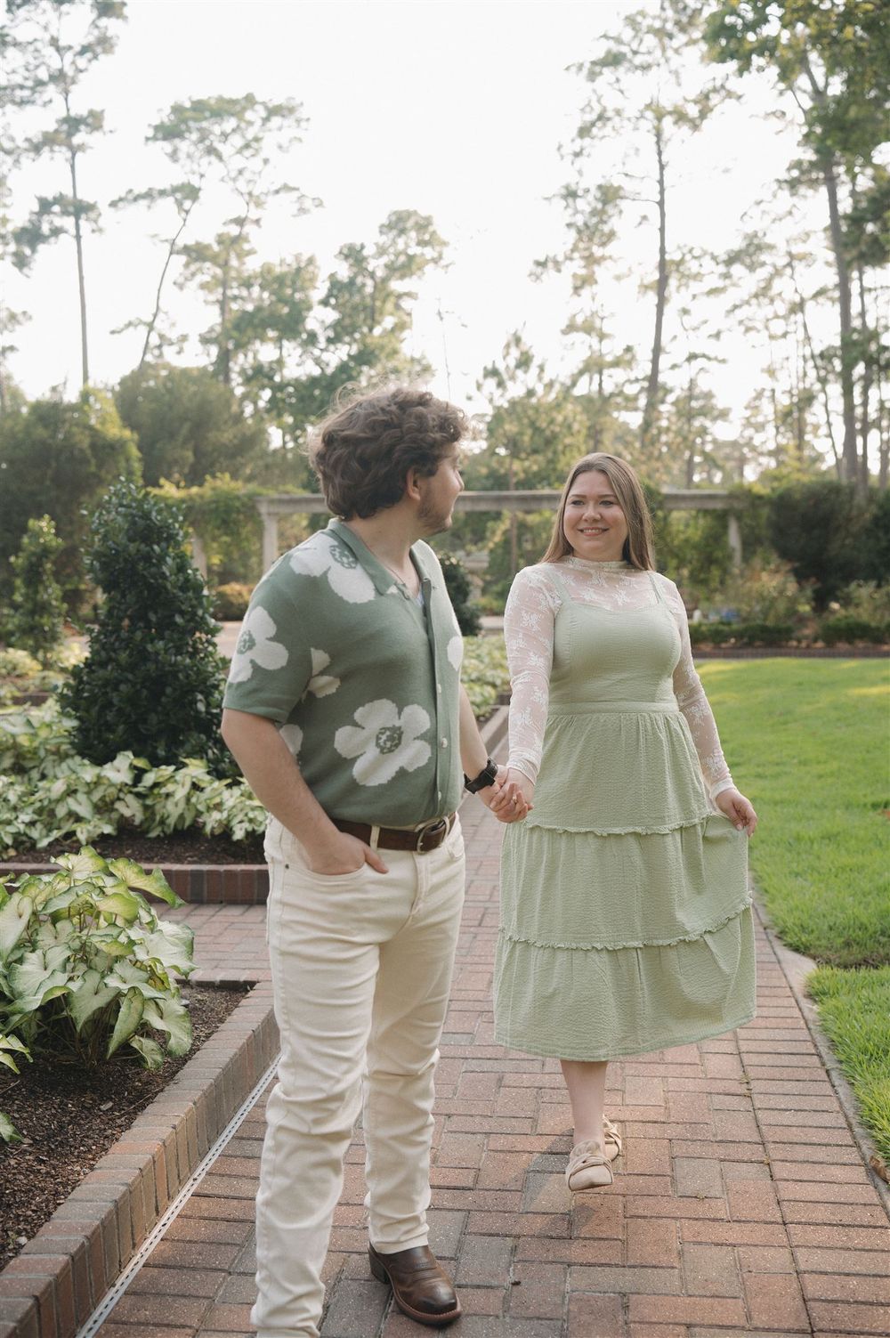 A couple walks hand in hand on a brick path in a garden setting, wearing coordinated green and white outfits.