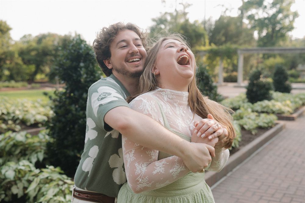 Two friends laughing and embracing together in a garden path surrounded by green shrubs and trees in warm sunlight.