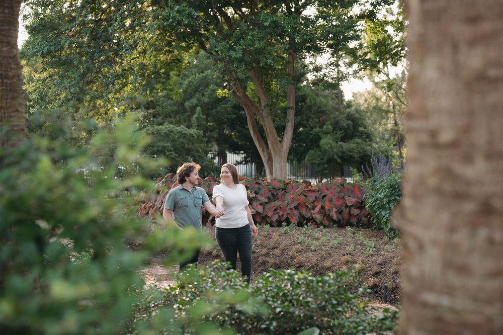 Two people walking together through a lush garden path with trees and pink flowering plants on a sunny afternoon.
