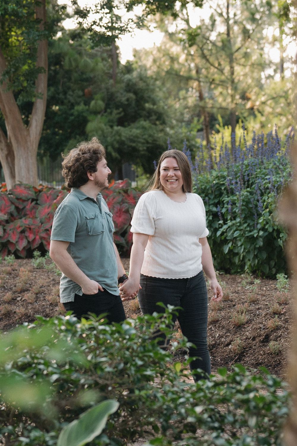 A couple shares a joyful moment while standing in a lush garden with purple flowers and greenery in the background.