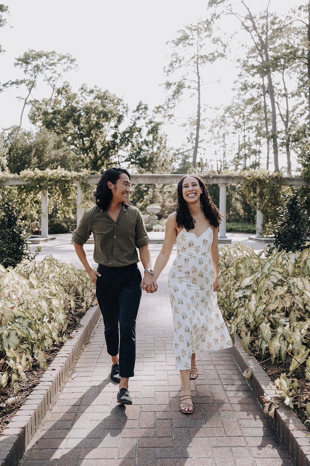 Couple walking hand in hand along a brick path surrounded by lush greenery and tall pine trees in a garden setting.