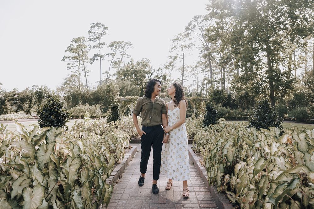 A couple walks hand in hand along a wooden pathway surrounded by lush green foliage and tall pine trees.