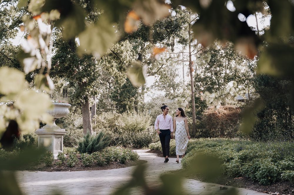 Two figures stroll together along a peaceful garden path framed by lush greenery and dappled sunlight.