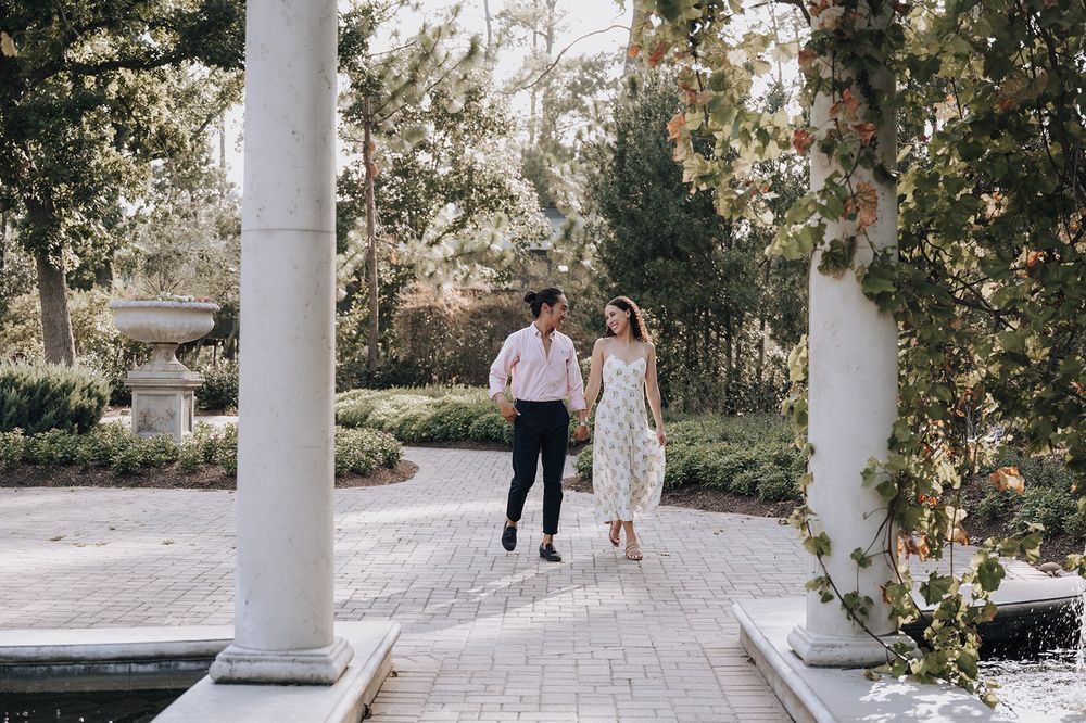 Couple walking together between white pillars along a paved garden path surrounded by lush greenery.