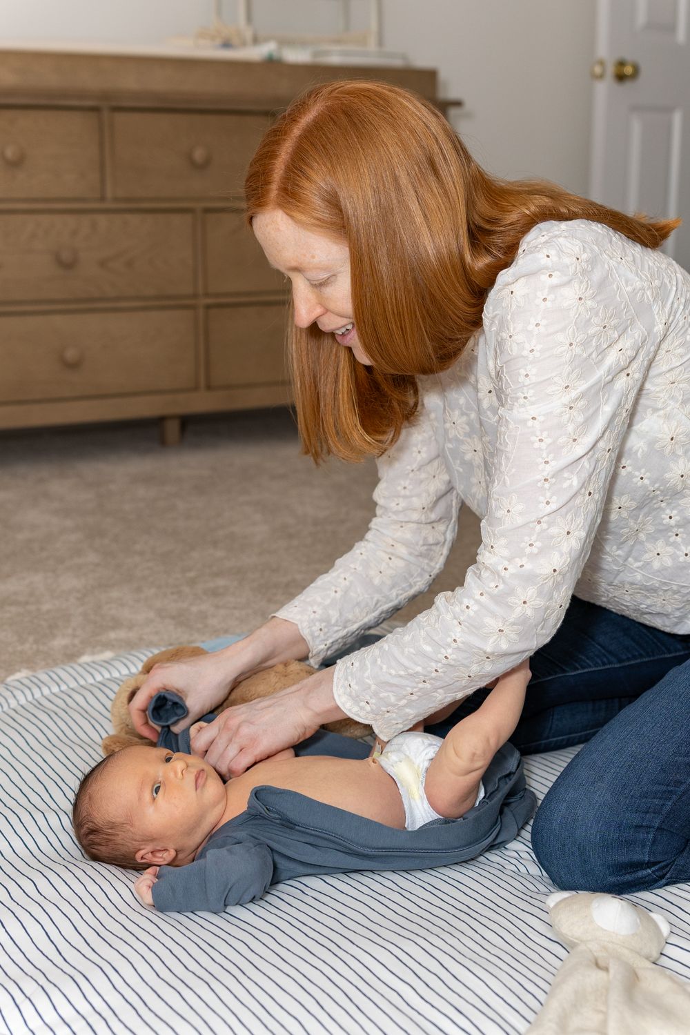 Parent dressing infant in blue clothing on striped bedding with wooden dresser in background during a newborn photo shoot in Sudbury, MA