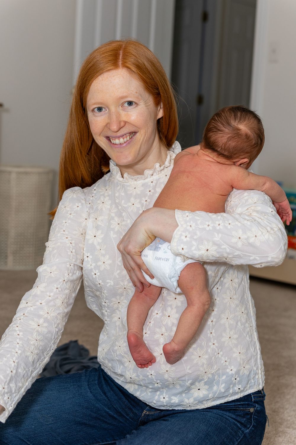 Woman in white eyelet blouse holds newborn baby indoors against neutral background during a newborn portrait shoot outside of Boston, MA