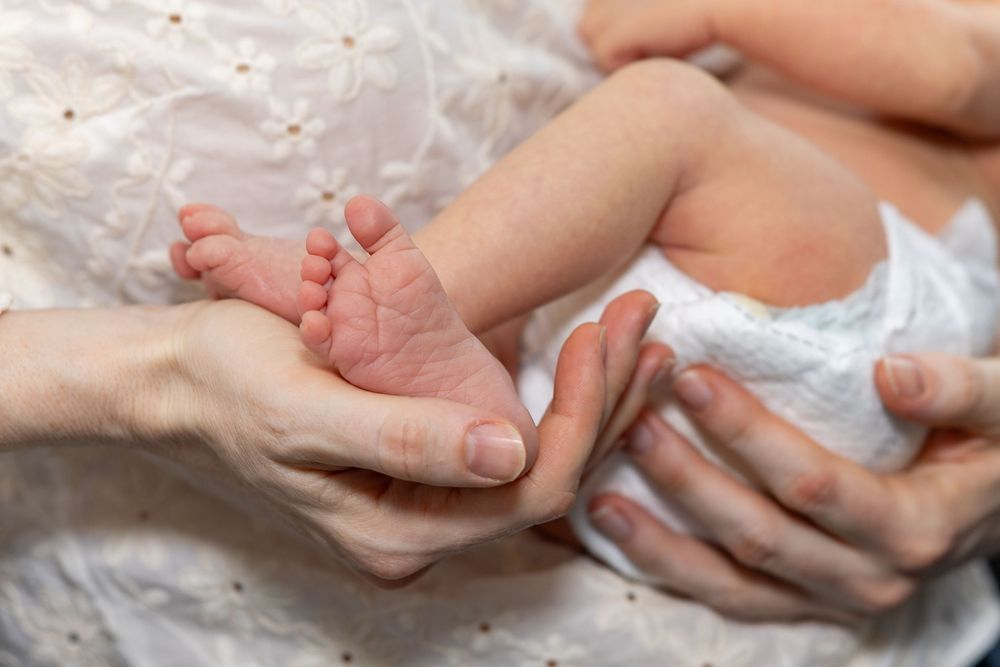 Gentle hands hold tiny newborn feet against white fabric background during a newborn portrait photo shoot in Sudbury, MA outside Boston, MA