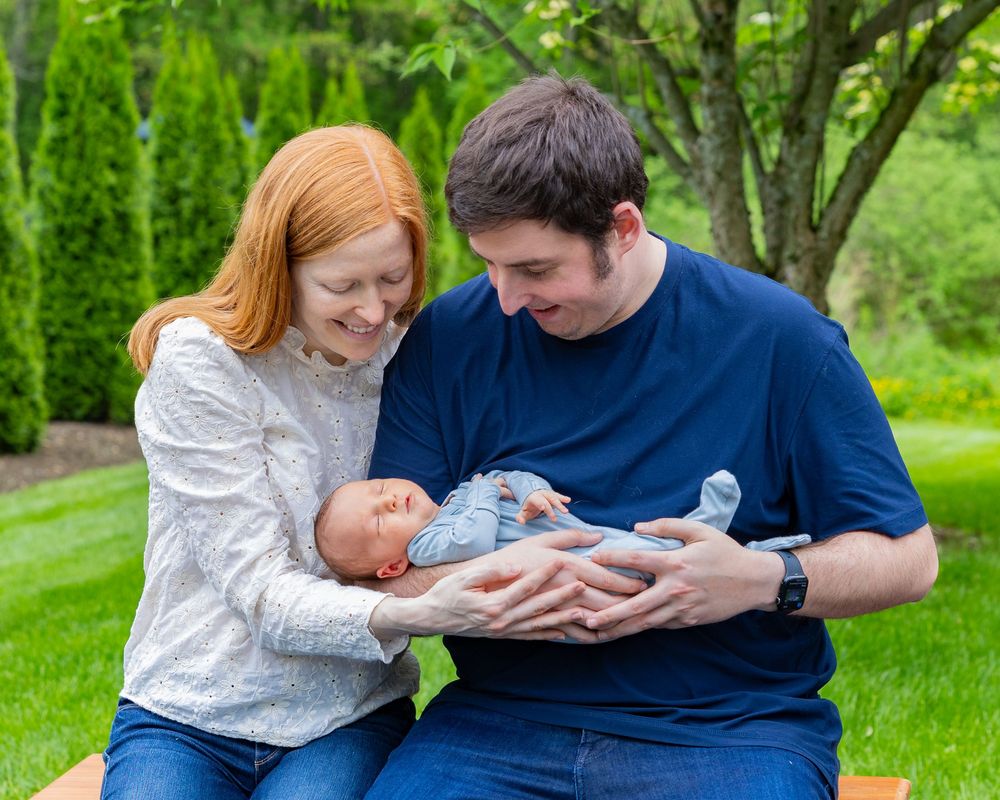 Couple sitting outdoors with their newborn baby dressed in light blue clothing during a newborn portrait photo shoot in Sudbury, MA.