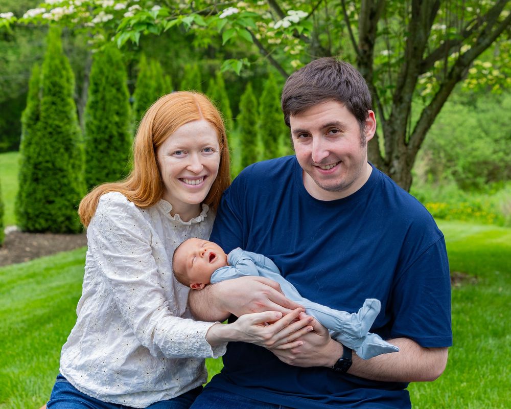 Happy parents holding their newborn baby while sitting together in a beautiful garden during a newborn portrait photo shoot in Sudbury, MA.
