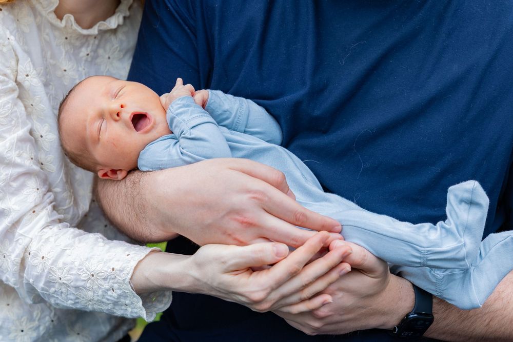 Close-up of yawning baby in blue outfit being held by parents' hands during a newborn portrait shoot outside of Boston, MA in Sudbury.