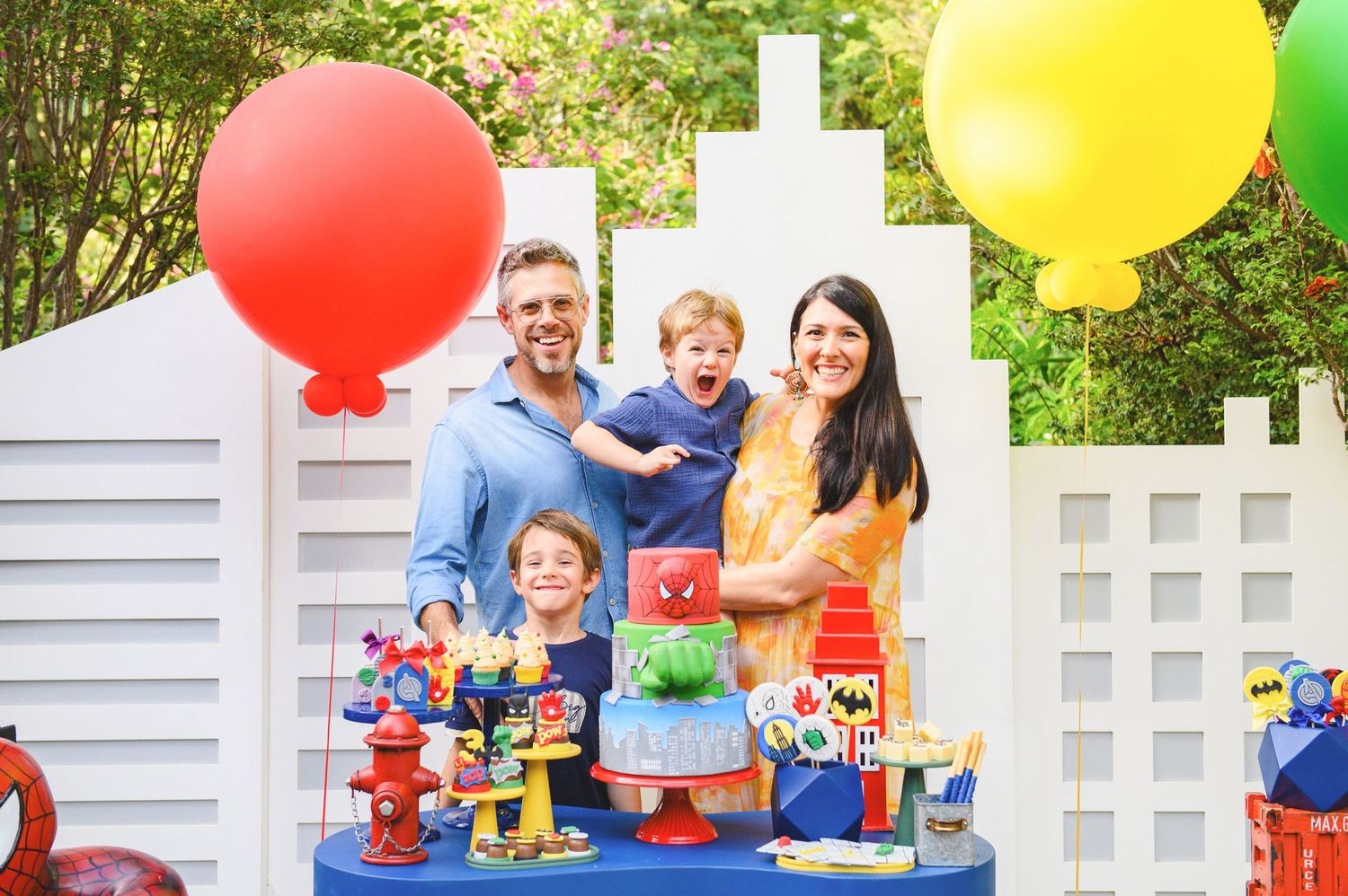 A festive birthday party setup with colorful balloons and toys on a blue table against a white fence outdoors.