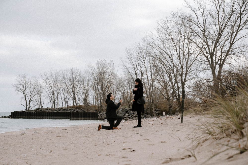A proposal moment captured at a beach with bare trees and overcast sky in the background.