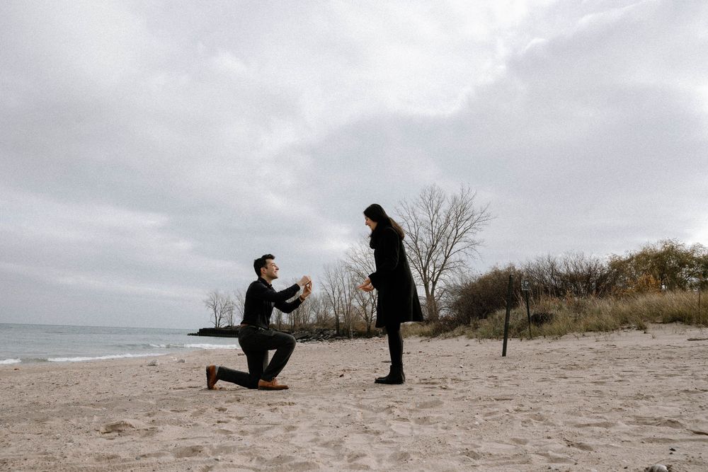A romantic beach proposal silhouette against cloudy skies with one person kneeling before another on sandy shores.