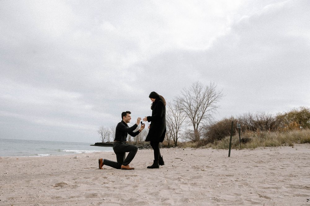 A romantic beach proposal moment captured on a cloudy day with bare trees and sand dunes in the background.