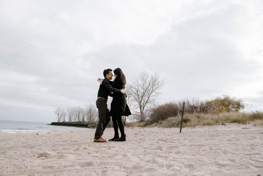 Couple in dark winter coats embrace on a cloudy beach with bare trees and sand dunes in the background.