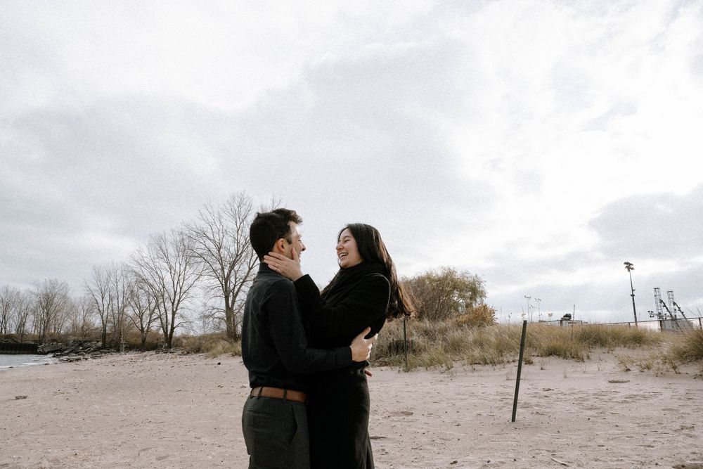 A couple embraces on a cloudy beach landscape with bare trees and sand in the background.