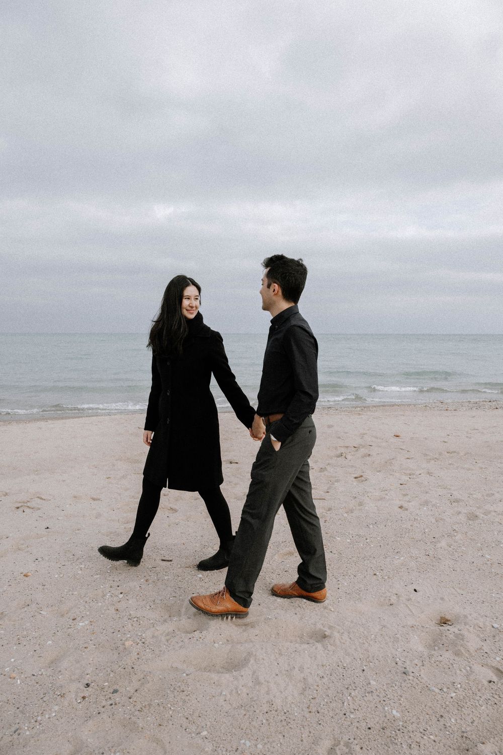 A couple holding hands while walking along a misty beach shore wearing black and grey outfits.