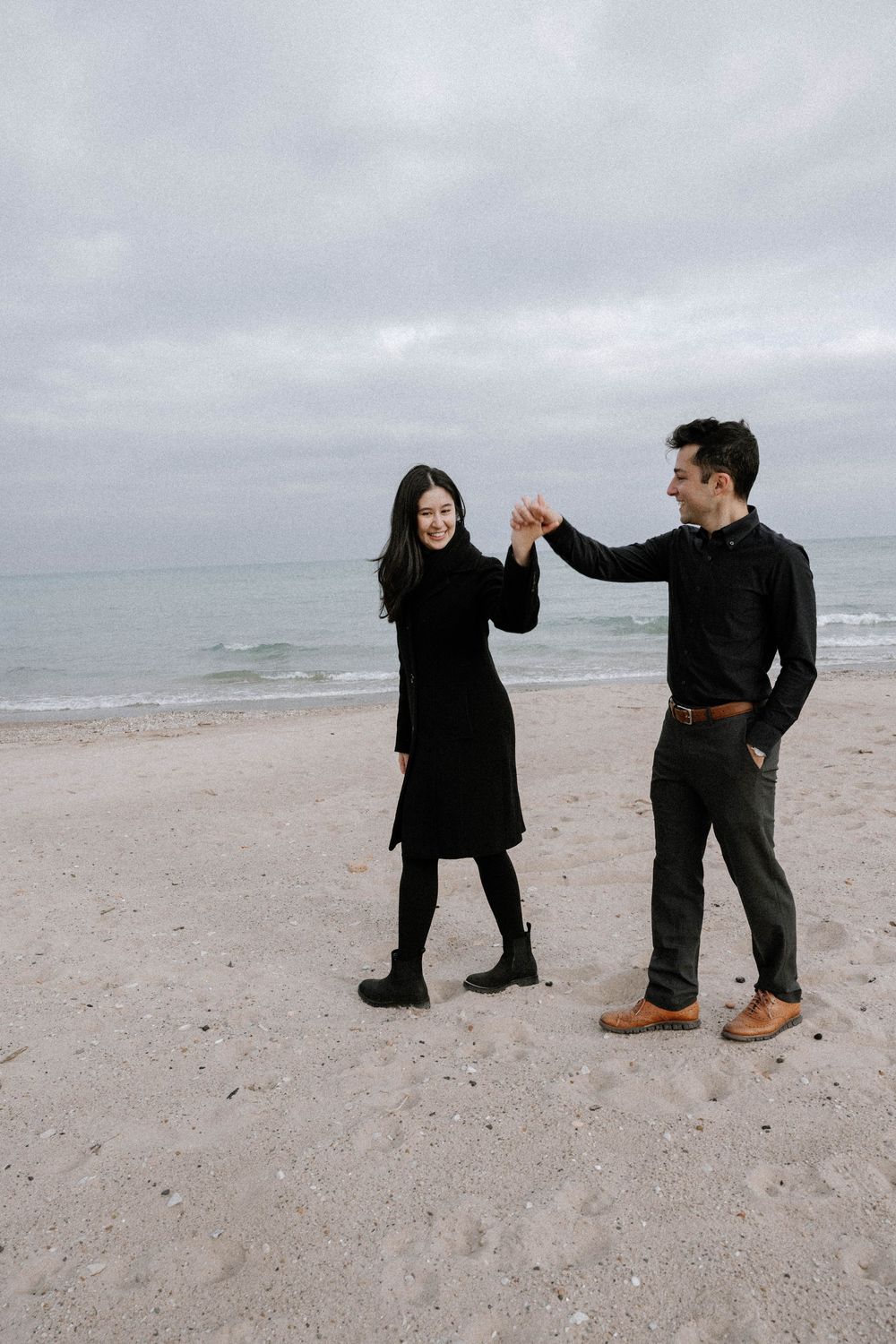 A couple in black outfits dancing together on a cloudy beach with the ocean waves visible in the background.