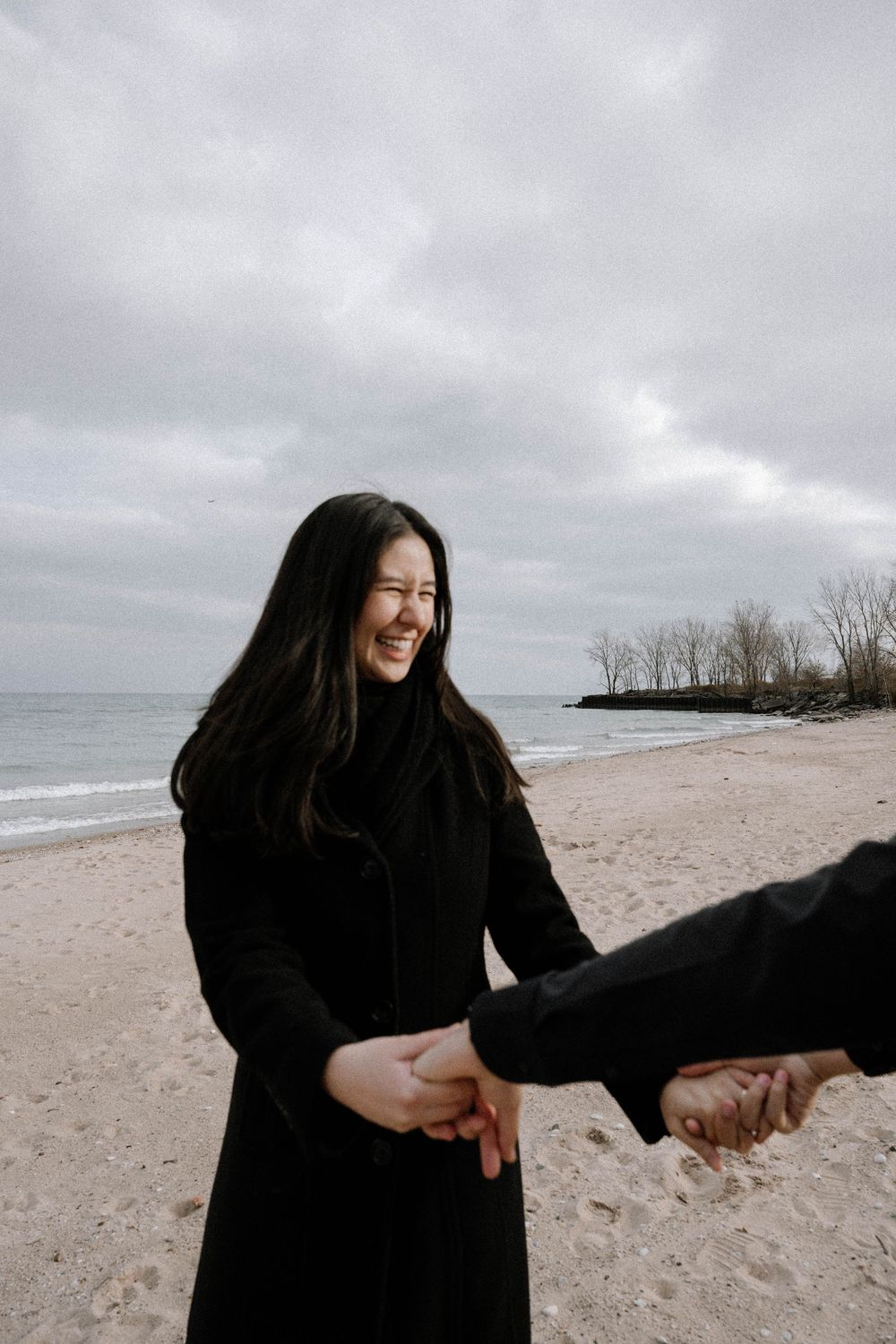 A couple holding hands on a cloudy beach with waves and bare trees in the distance.