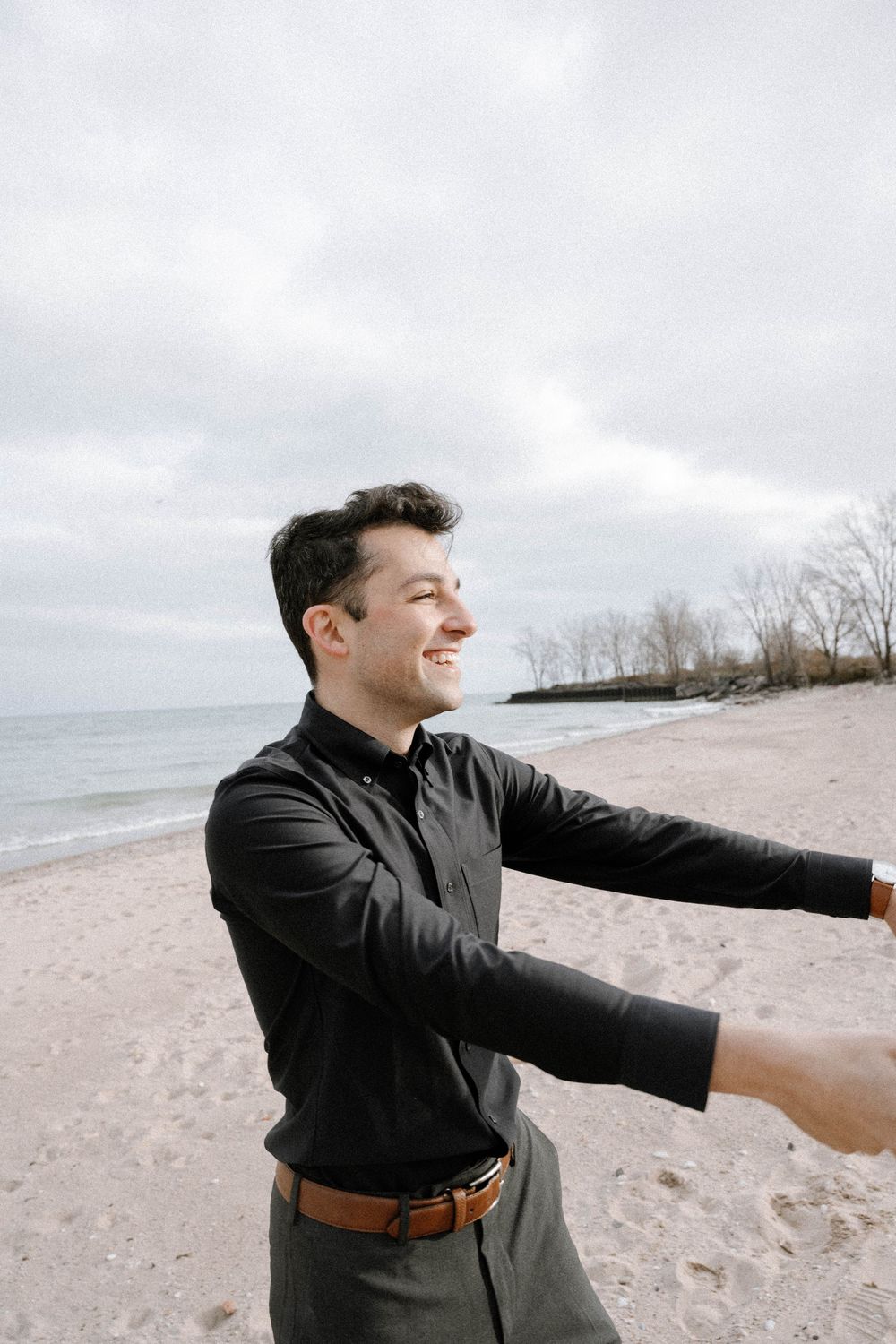 Person in dark clothing smiling and extending arms outward while standing on a cloudy beach by the water.