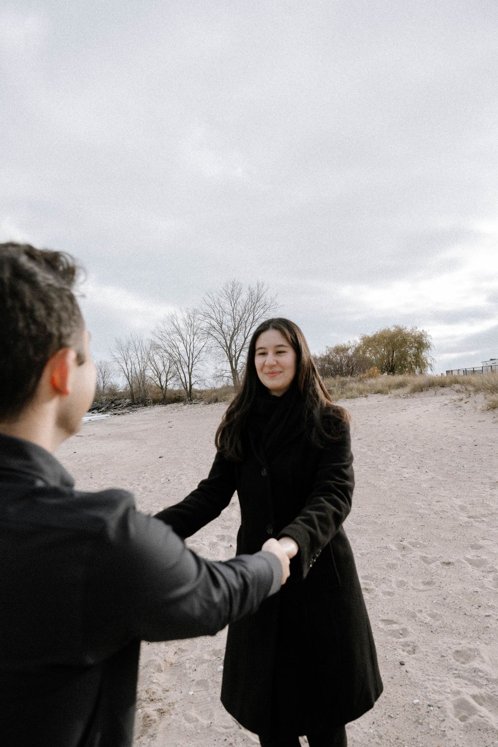 Two people in dark winter coats hold hands on a sandy beach under overcast skies.