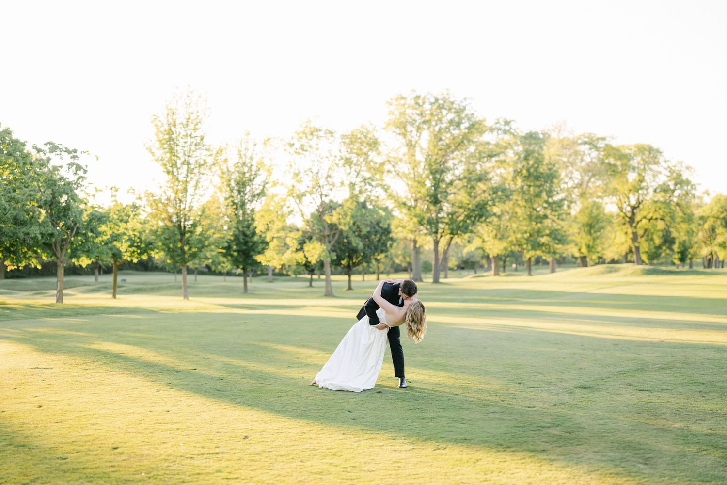 A romantic wedding photo of a couple embracing on a lush green golf course in golden afternoon sunlight.