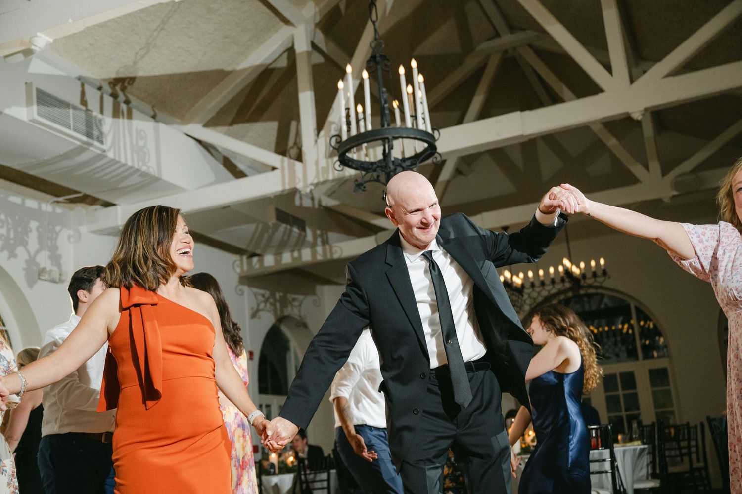 Wedding guests dancing and celebrating on a wooden dance floor in an elegant reception venue with arched ceilings and chandelier.