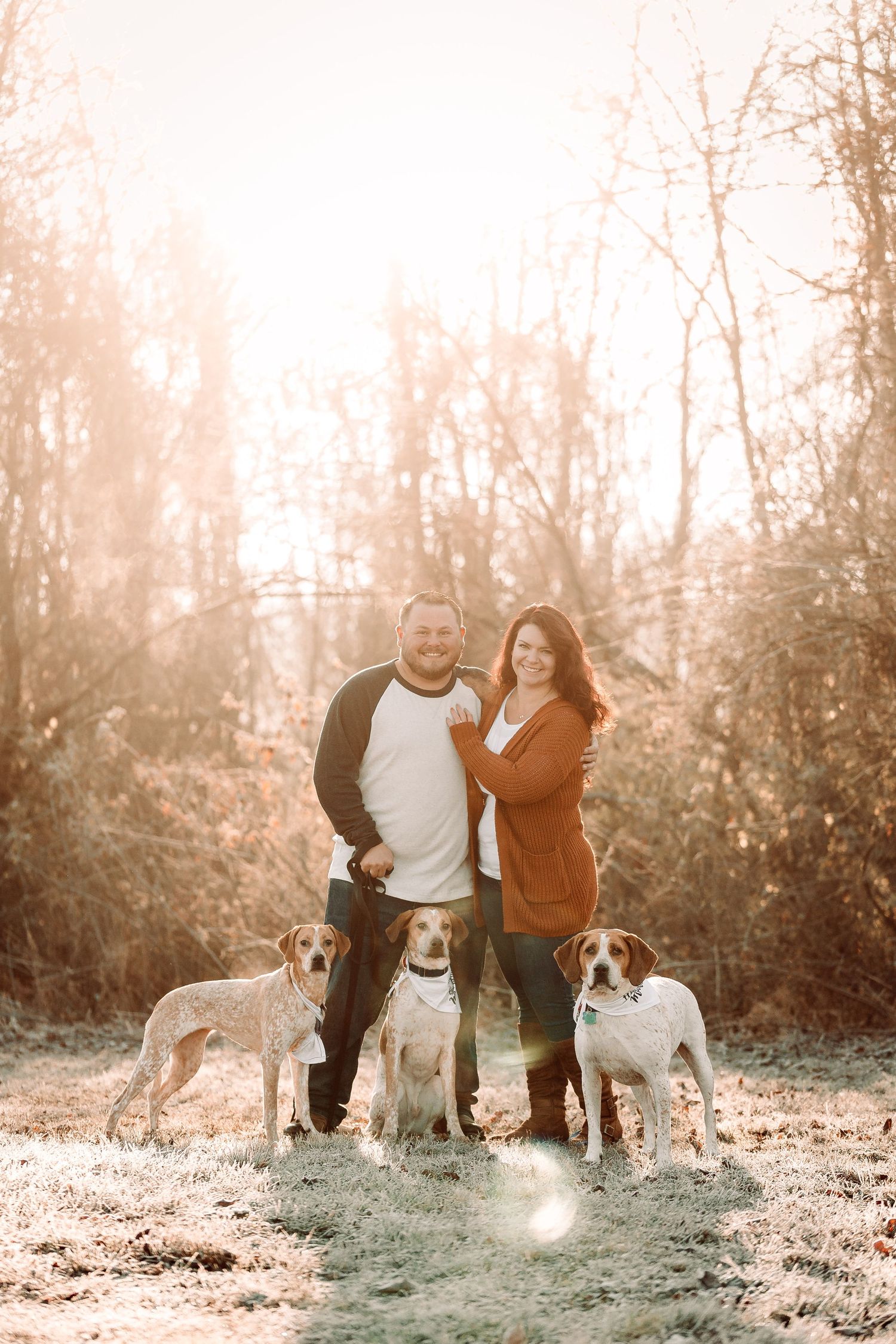 A couple stands outdoors in warm sunlight with their three dogs on a frosty morning in the woods.