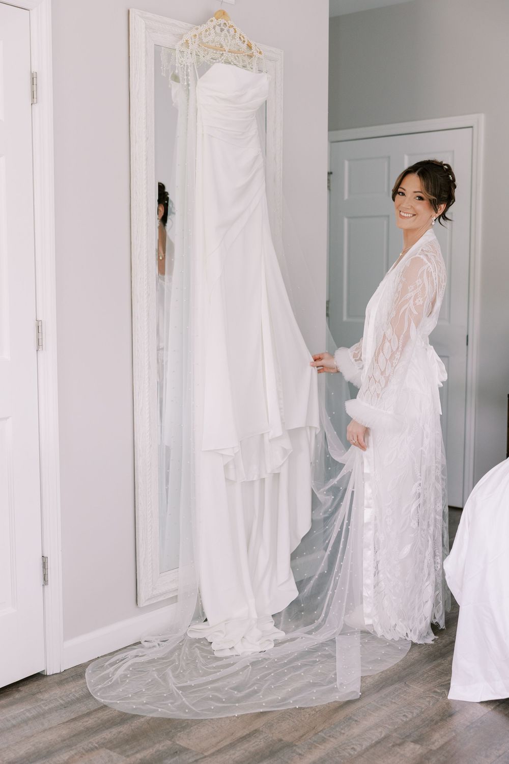 A delicate white lace robe-clad person admires an elegant wedding gown with a long train displayed on an ornate mirror at Windham Manor.