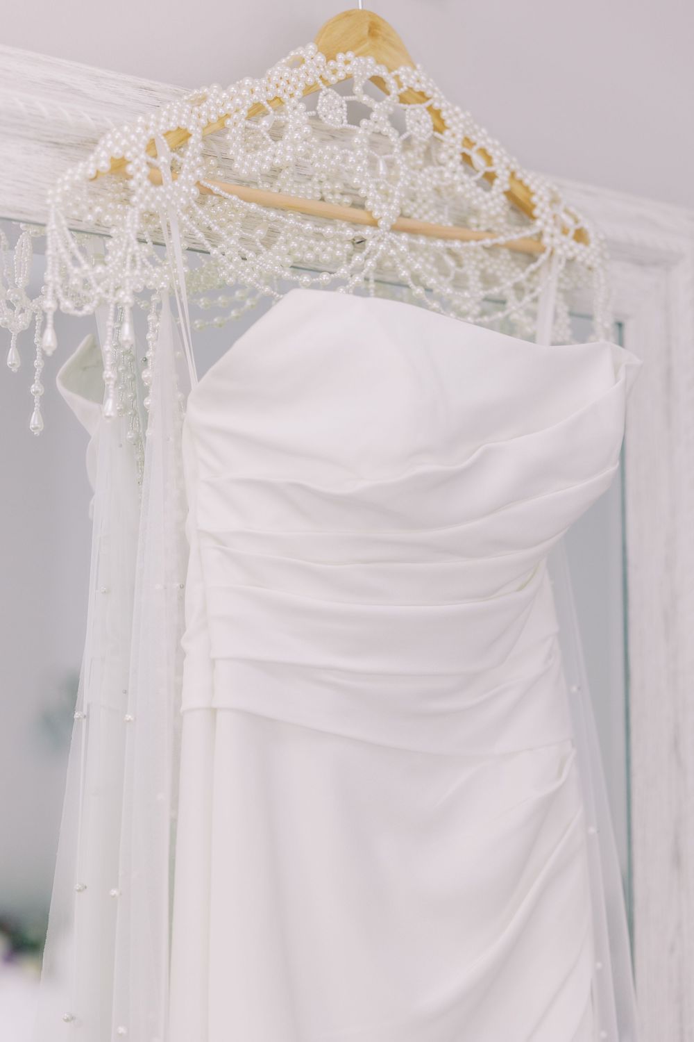 A white wedding dress with ruched detailing hangs on a beaded hanger with intricate lace details at Windham Manor.