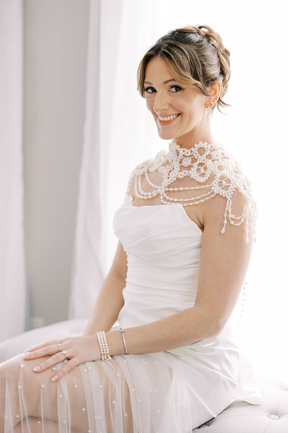 A bride in an elegant white wedding dress with delicate lace capelet sits smiling while wearing pearl bracelets at Windham Manor.