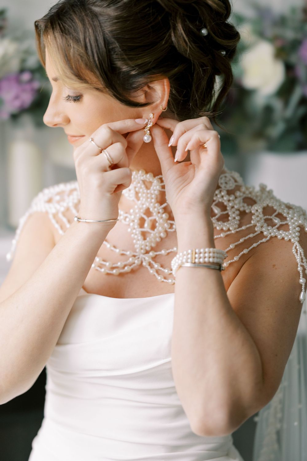 A bride putting on pearl earrings wears a delicate lace wedding dress with pearl jewelry and an updo hairstyle at Windham Manor.