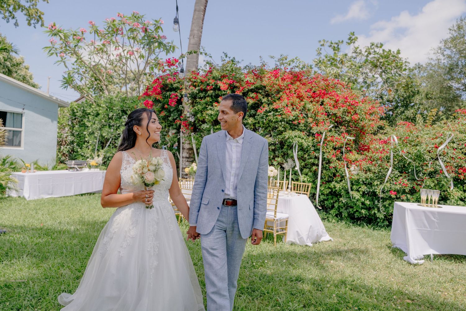 A formal outdoor wedding celebration under a floral archway with red bougainvillea flowers on a sunny day.