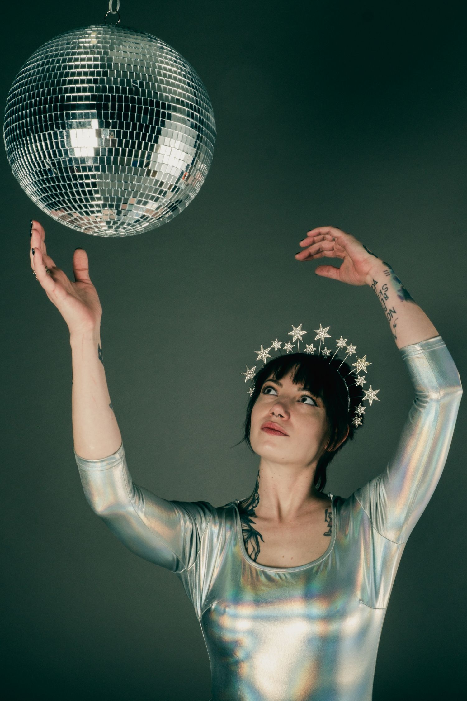 A dancer in a metallic silver dress and star crown poses with a disco ball against a dark green background.
