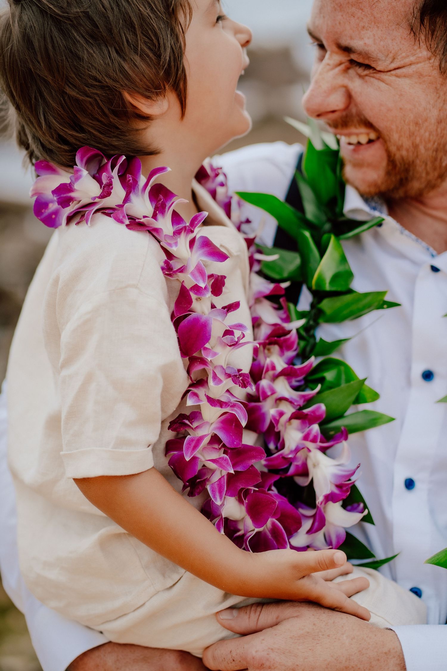 A laughing young child wearing a white shirt and vibrant purple and white orchid lei shares a joyful moment with an adult.
