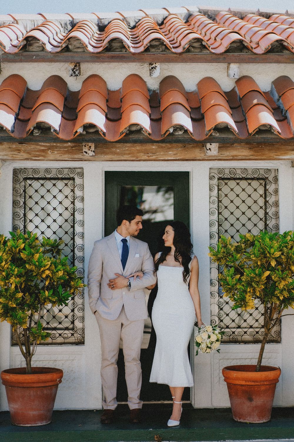 A couple stands in the doorway of a Spanish Colonial style building with terracotta roof tiles and potted plants.