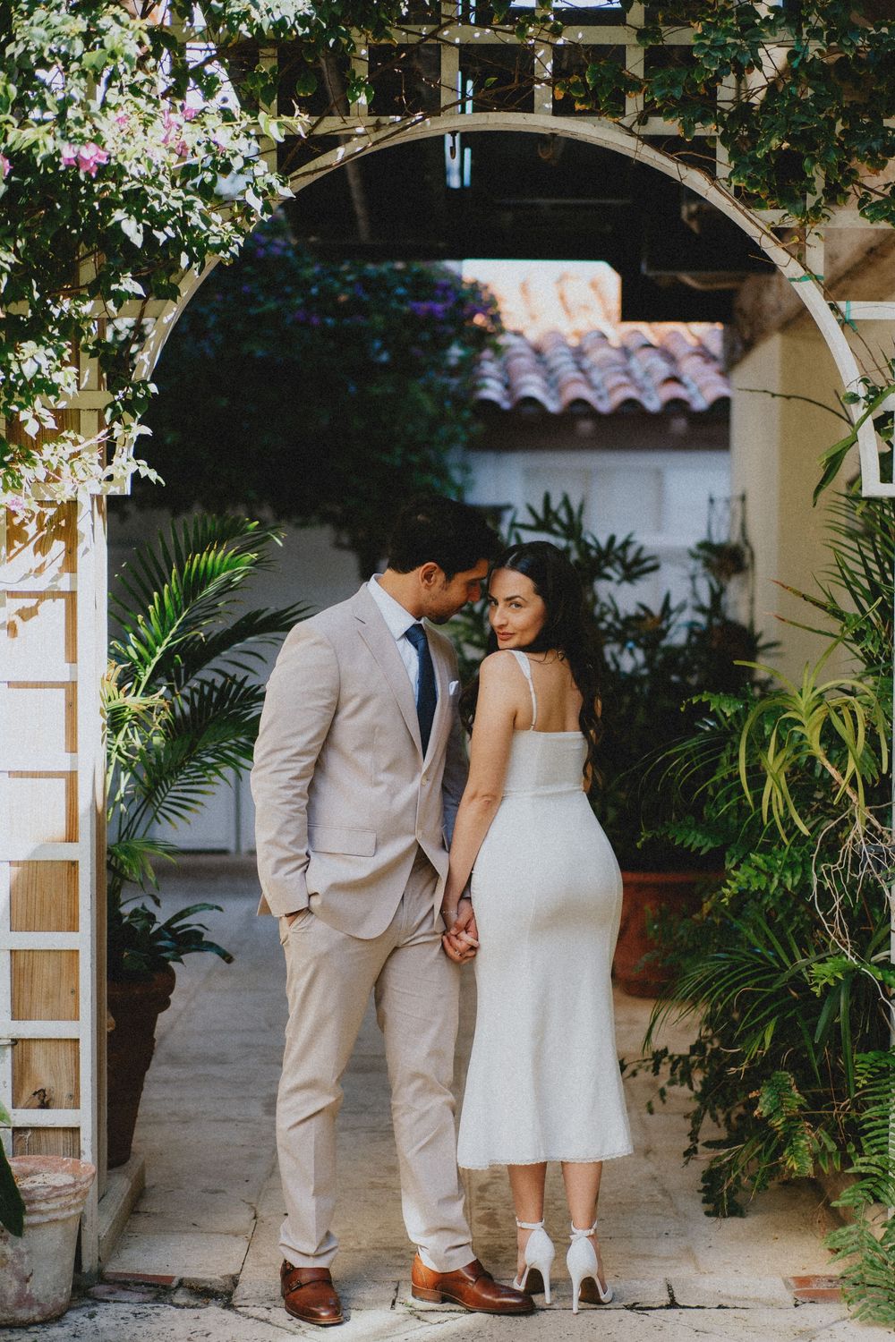 Romantic couple in elegant attire share an intimate moment under a Mediterranean archway surrounded by greenery.