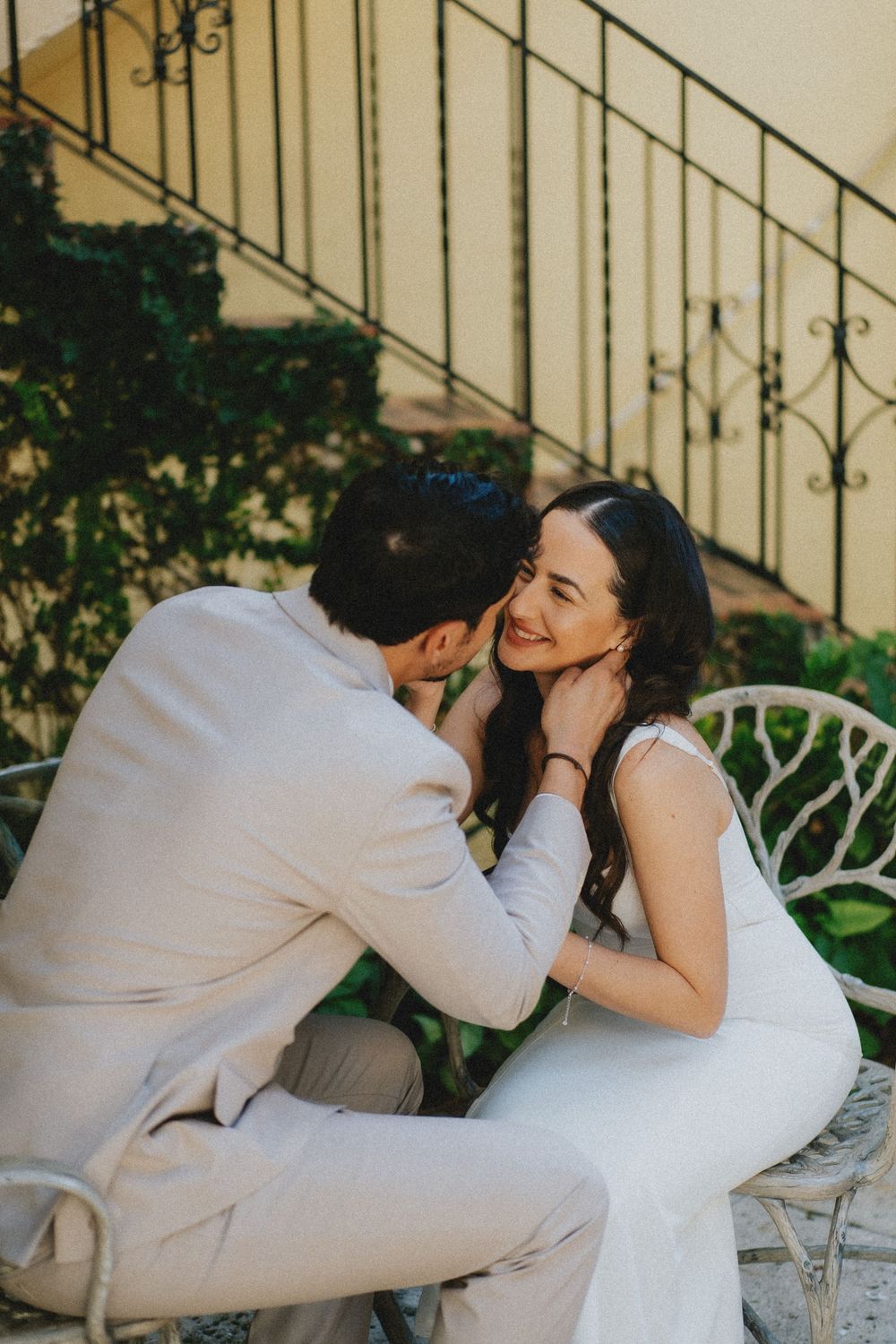 A couple in white outfits share an intimate moment while seated outdoors near wrought iron stairs.